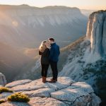 A couple stands on a rocky cliff overlooking a vast, rugged landscape. The sun sets behind them, casting a warm glow over the scene