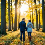 A couple stands in a colorful autumn forest, surrounded by fallen leaves and warm sunlight filtering through the trees